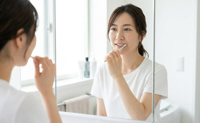 鏡に映る自分の顔を見ながら、穏やかに手元で歯を磨く日本人女性の実写画像。
