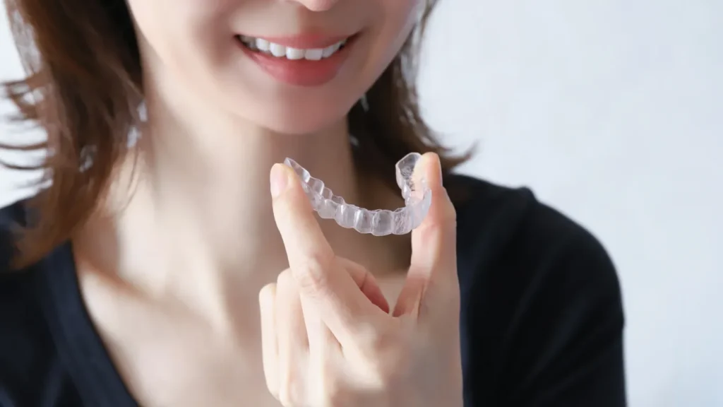 透明なマウスピース型矯正装置を指先でつまんで見せる女性（黒いトップス）