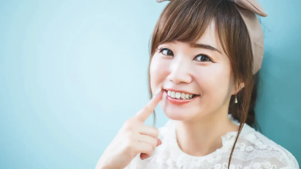 水色の背景の前で人差し指を自分の前歯に添えて笑う女性（顔と口元の見え方）